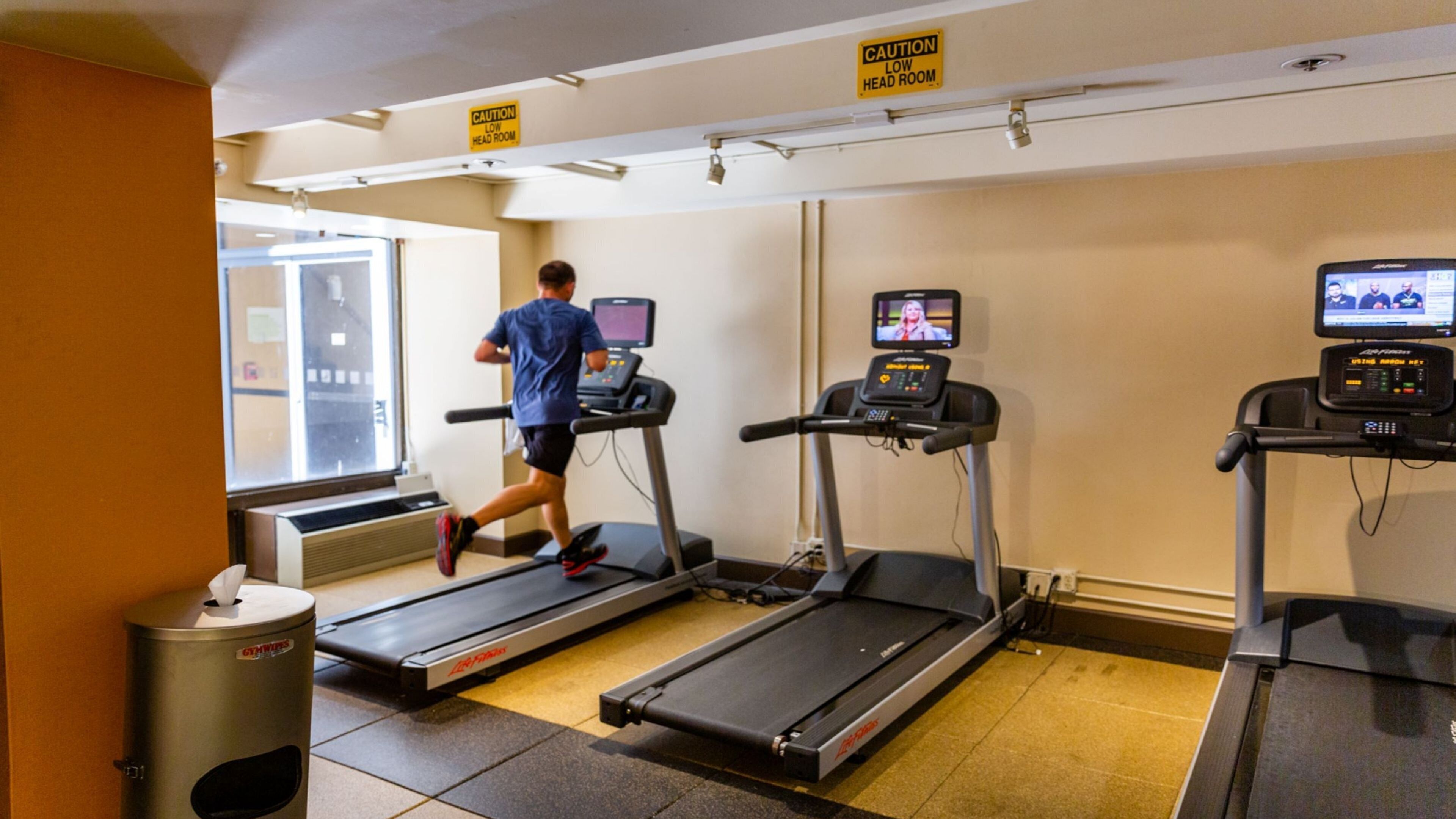 A fitness room with treadmills, a caution sign for low head room, and a window with a view.
