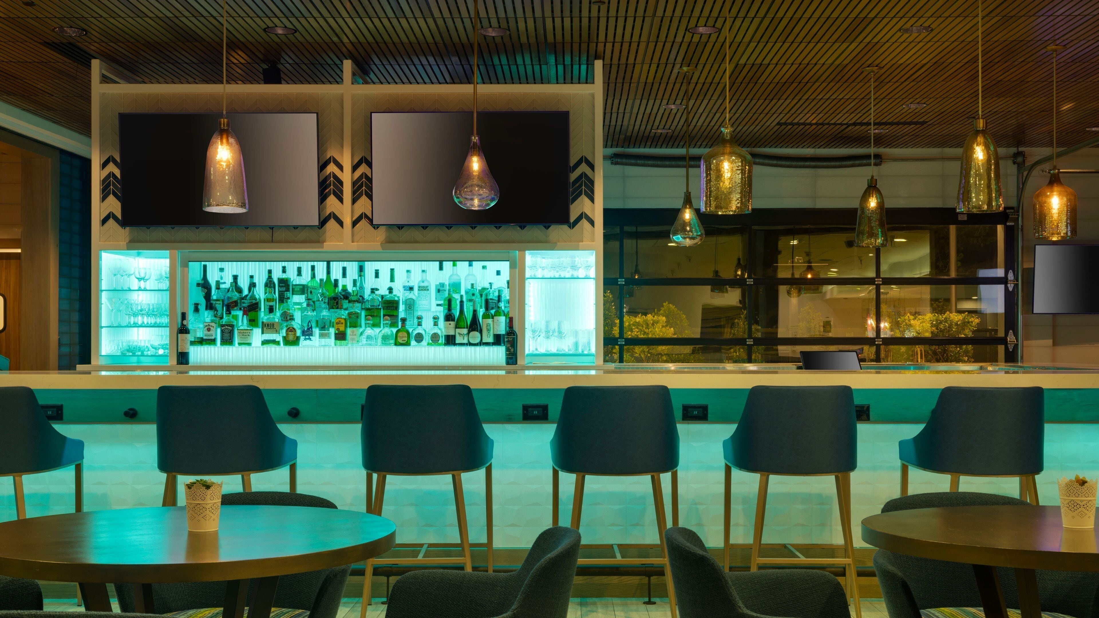 A bar area with a modern design, featuring a bar counter with a variety of bottles and glasses, and several chairs and tables for seating.