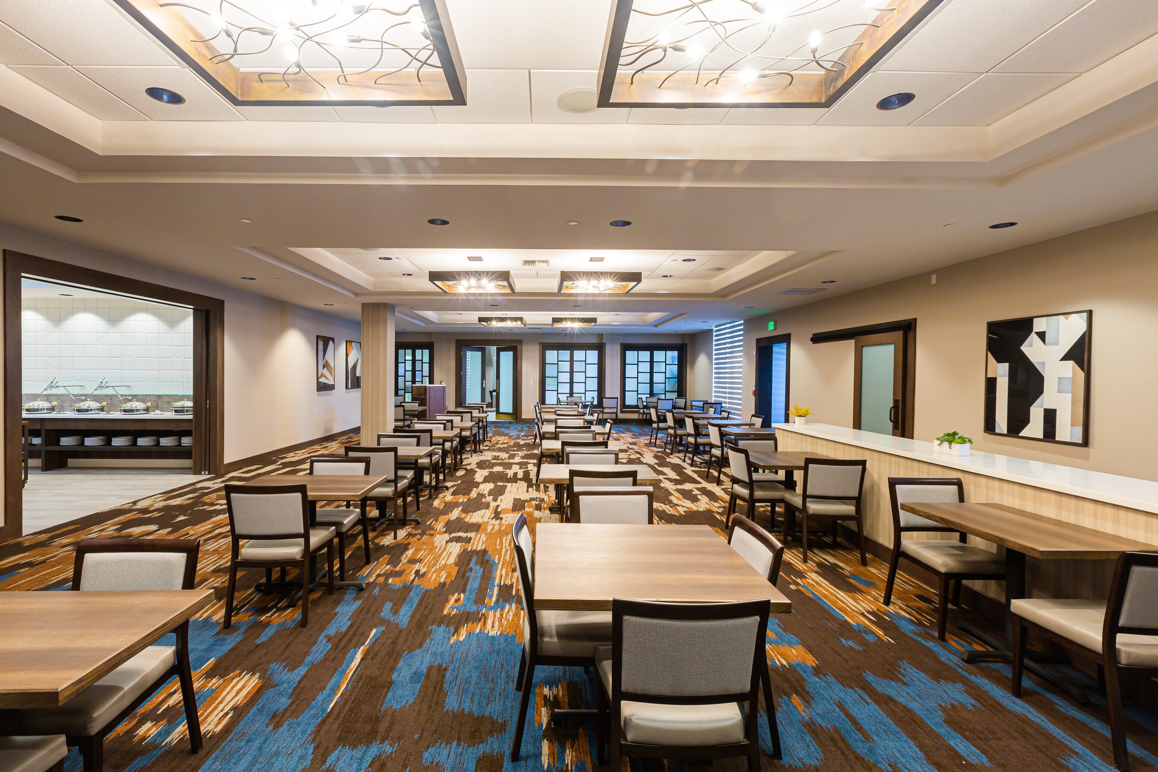 A spacious dining area with multiple tables and chairs, large windows, and a patterned carpet.