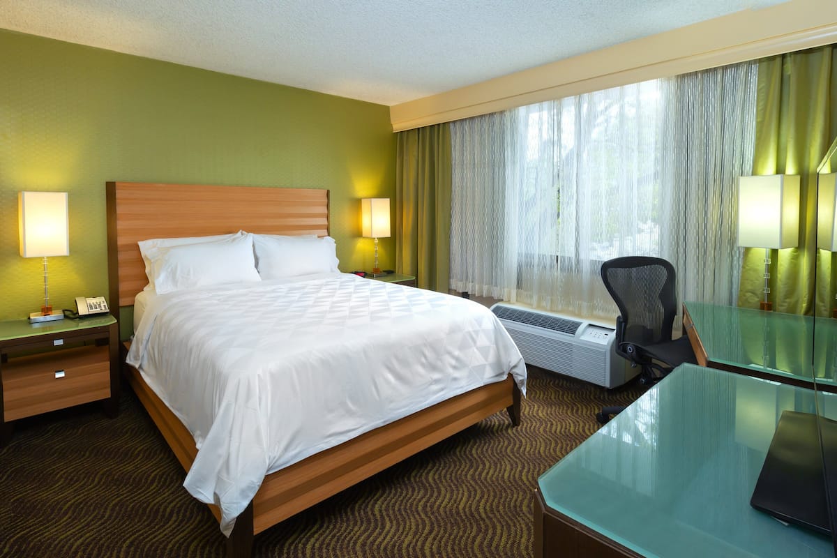 A hotel room with a large bed, two bedside tables with lamps, a desk with a chair, and a large window with curtains.