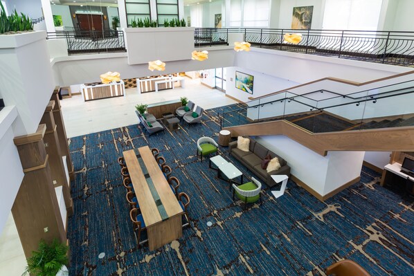 A spacious lobby with a high ceiling, a central seating area with sofas and chairs, a long wooden table, and a staircase leading to an upper level. - Holiday Inn San Jose - Silicon Valley by IHG (San Jose)