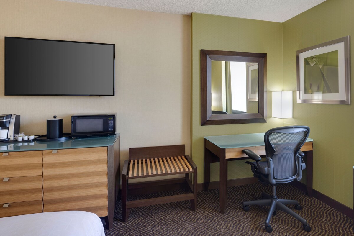 A hotel room with a bed, a desk with a chair, a television, a coffee maker, and a mirror.
