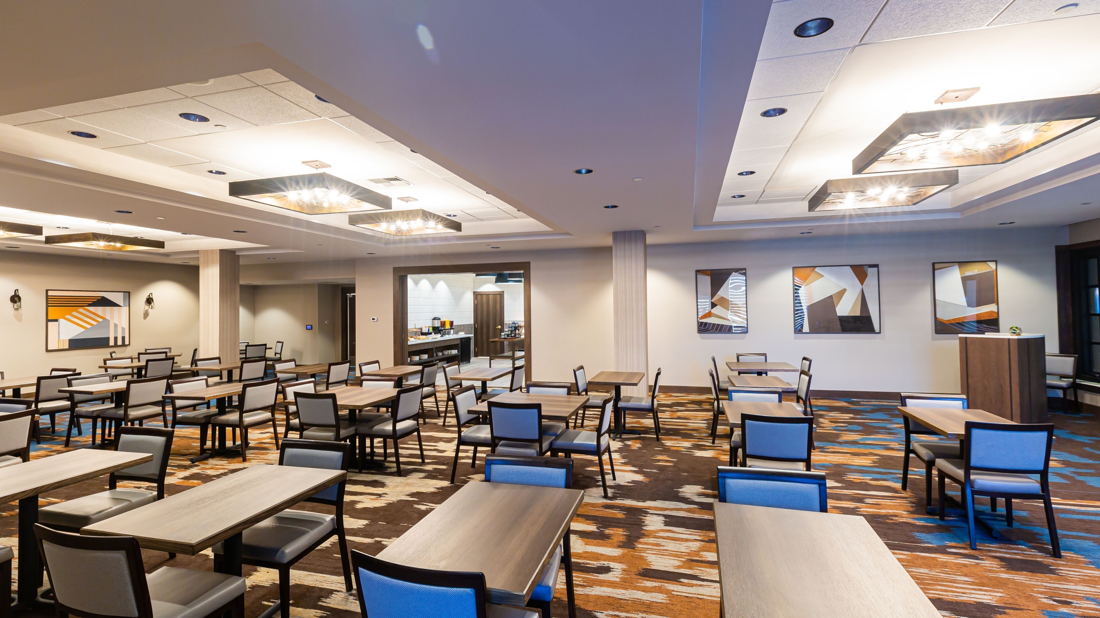 A hotel conference room with multiple tables and chairs, a kitchen area, and artwork on the walls.
