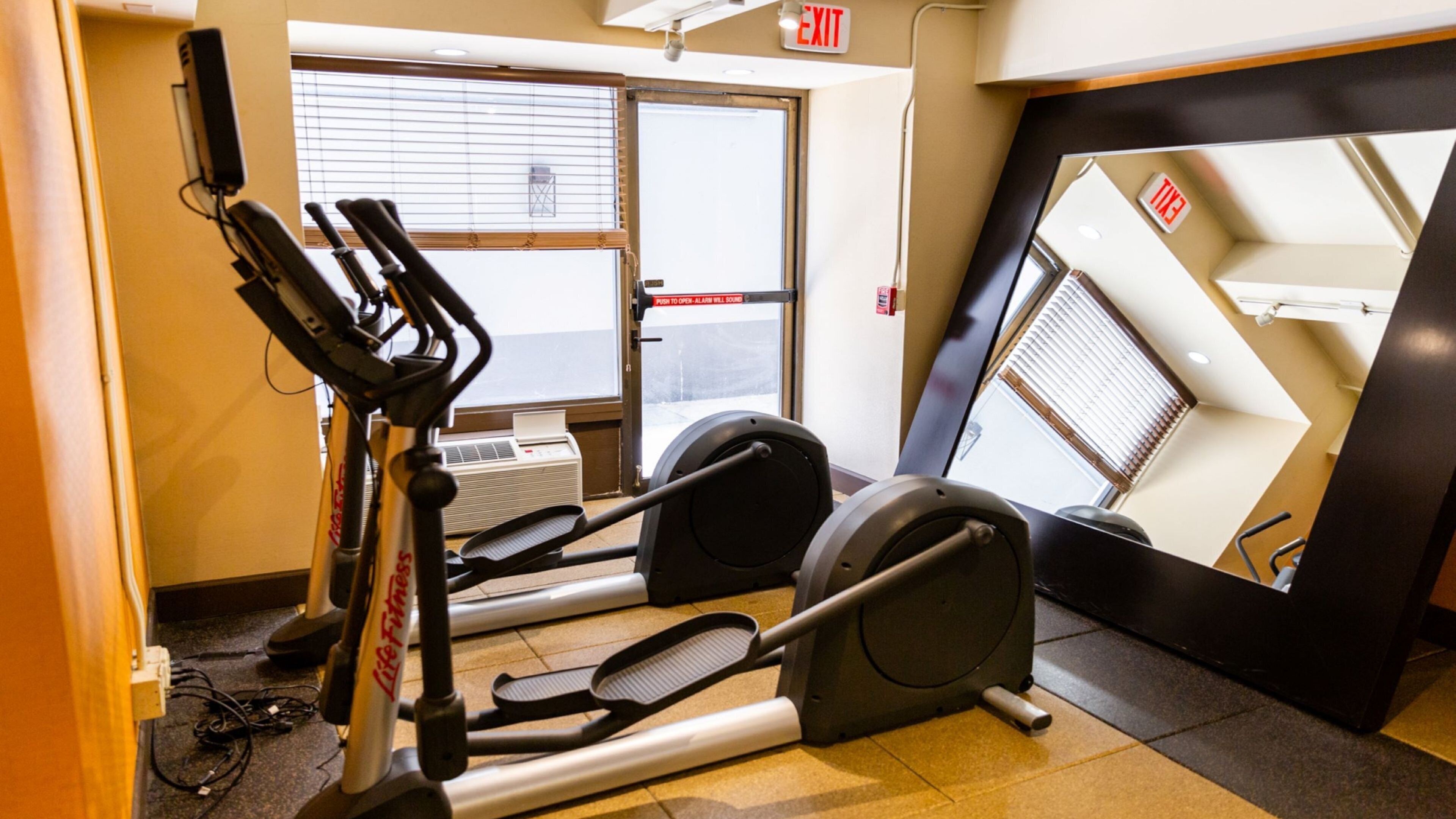 Un gimnasio con cintas de correr y una bicicleta estática, una ventana con persianas y una señal de salida.