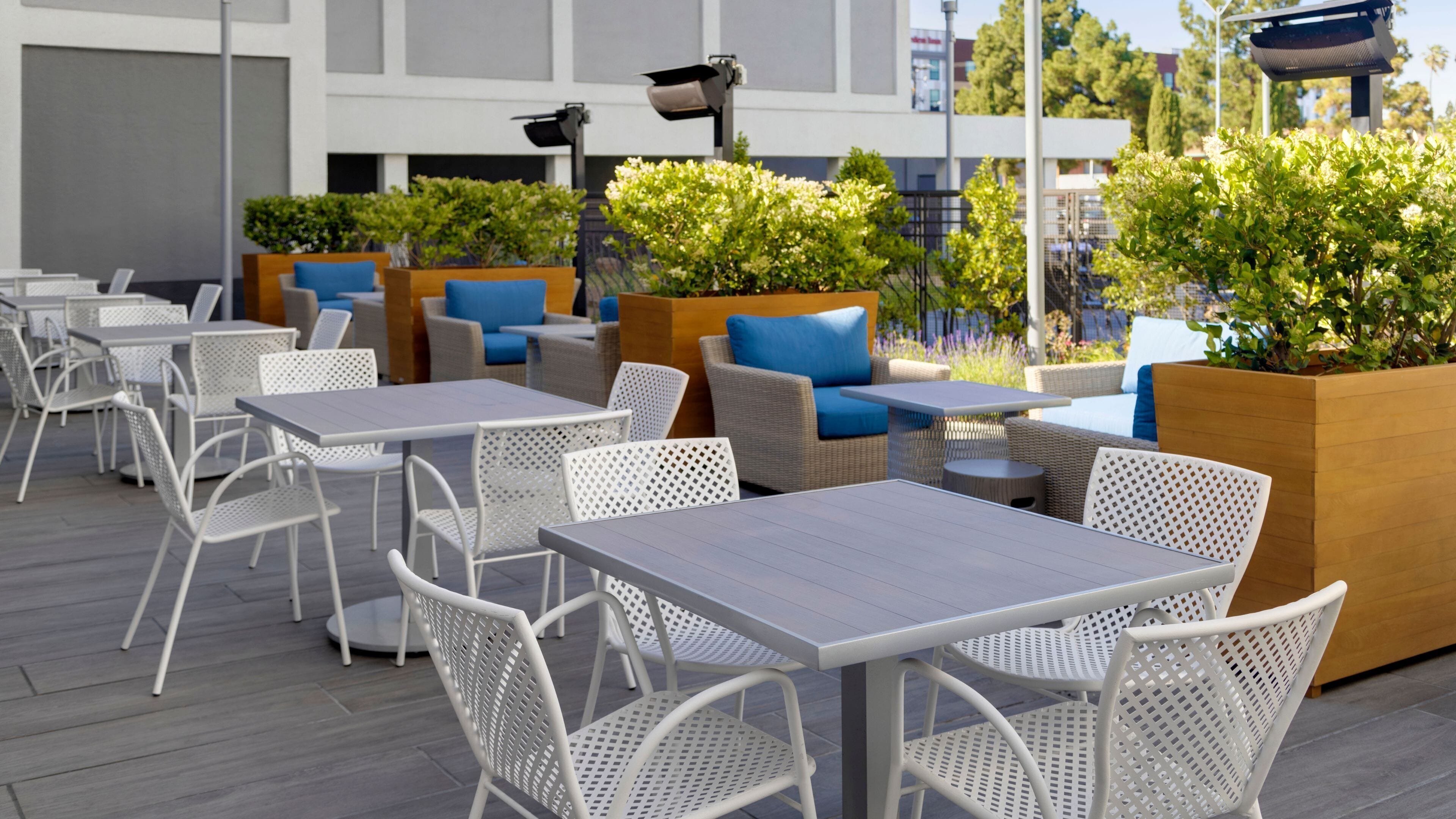 An outdoor seating area with white metal tables and chairs, wooden planters with greenery, and a modern building in the background.