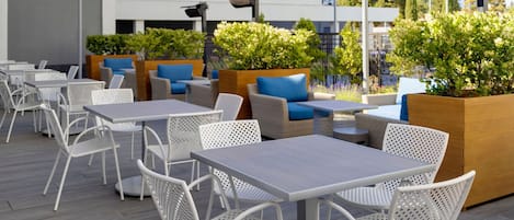 Een buitenzitplaats met witte metalen tafels en stoelen, houten plantenbakken met groen, en een modern gebouw op de achtergrond.