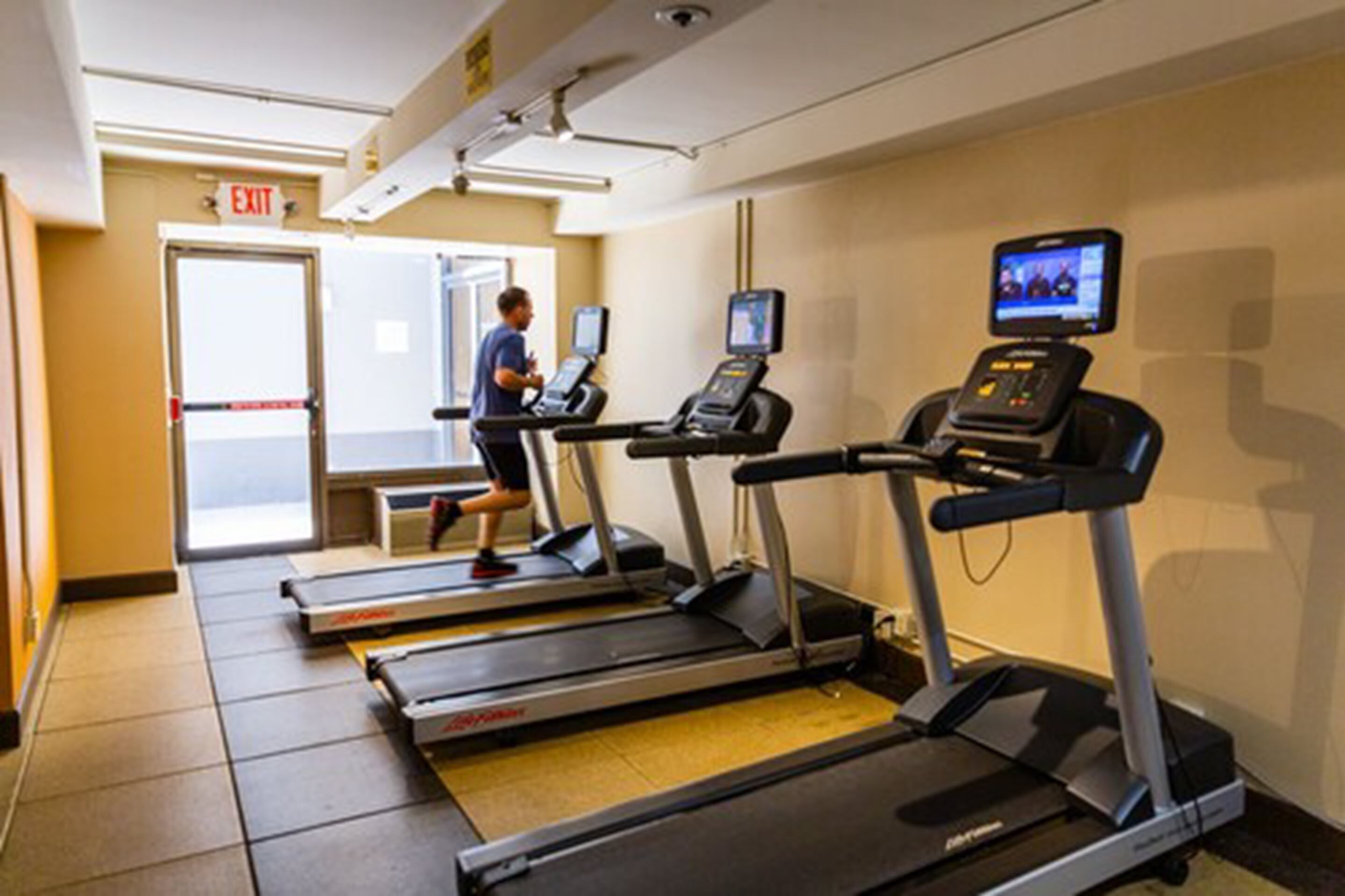 Une salle de remise en forme équipée de tapis de course, d’un écran numérique et d’un panneau indiquant la sortie.