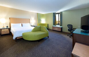 Una habitación de hotel con una cama, un sillón verde, un escritorio con una computadora y una televisión.
