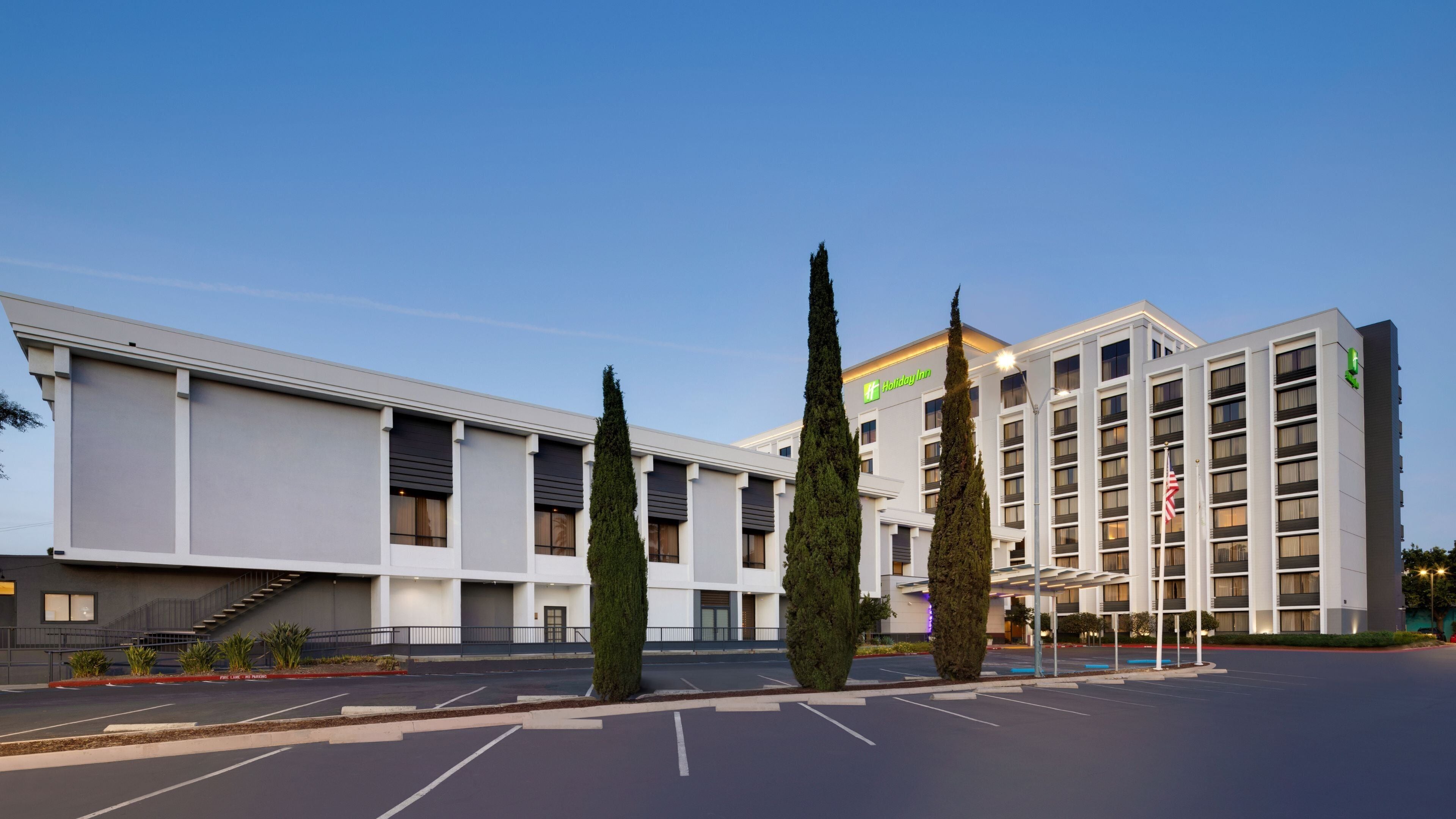 Un hotel Holiday Inn con diseño moderno, que cuenta con un estacionamiento y dos cipreses destacados.