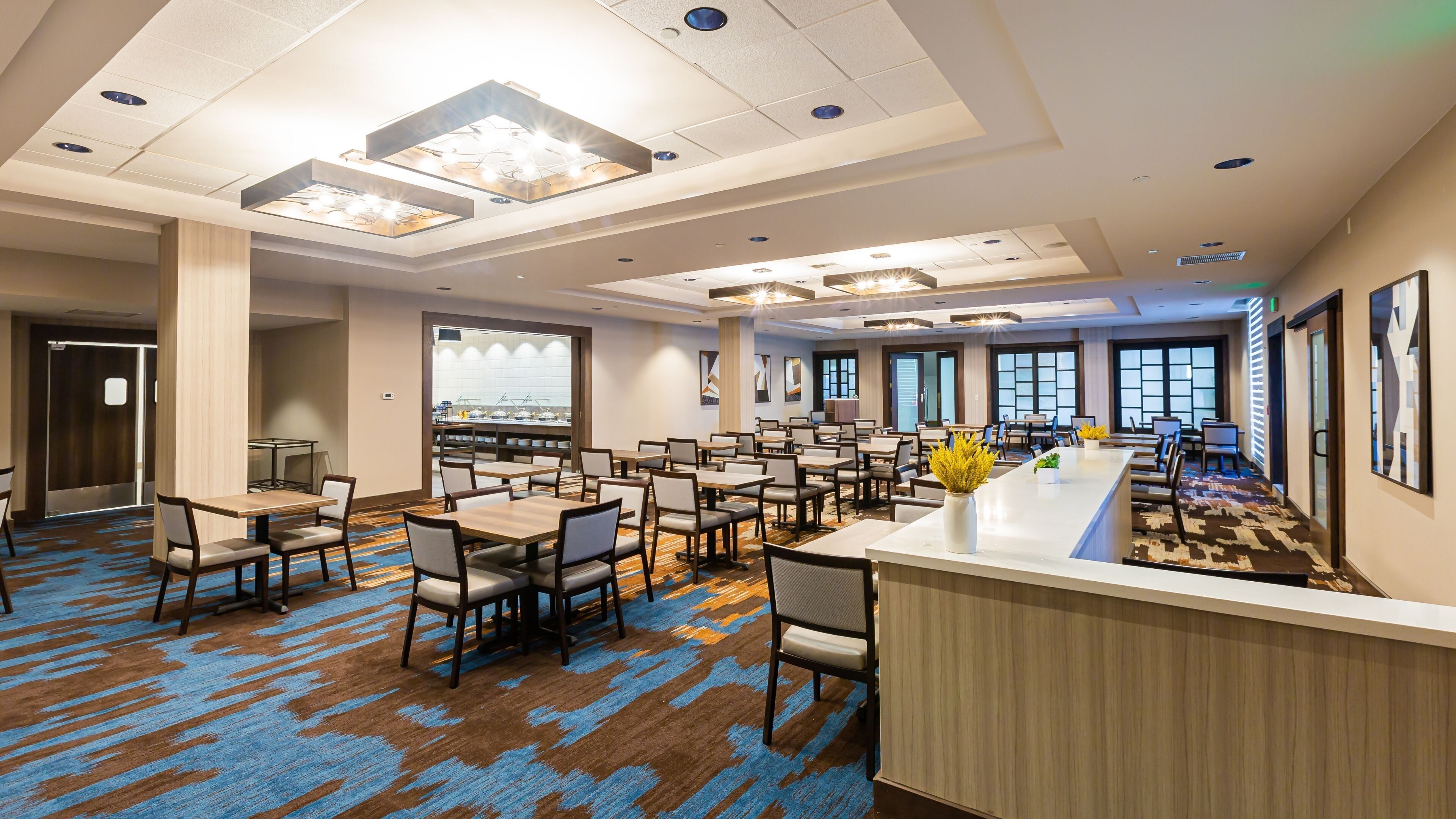 A hotel dining area with multiple tables and chairs, a central counter with a vase of flowers, and large windows providing natural light.