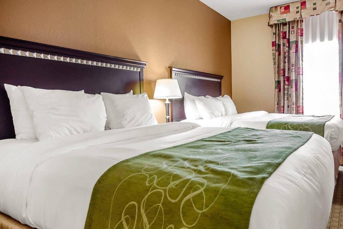 Ett hotellrum med två sängar, en sänggavel med ett dekorativt mönster, en lampa och ett fönster med gardiner.