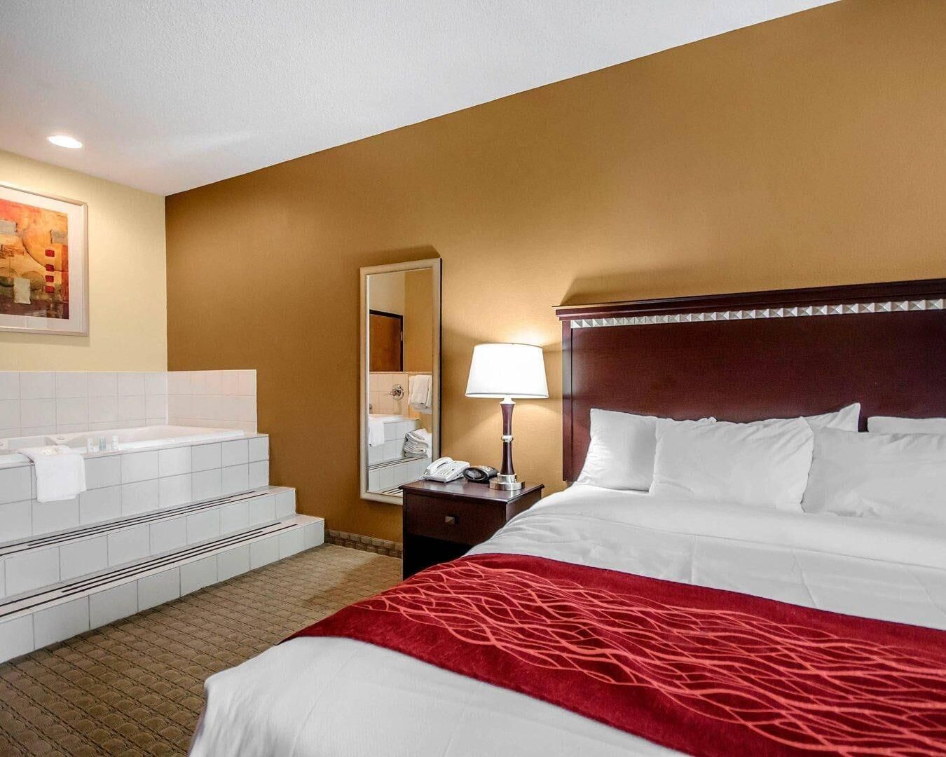 A hotel room with a bed, a nightstand, a lamp, a mirror, a bathroom with a bathtub, and a tiled floor.