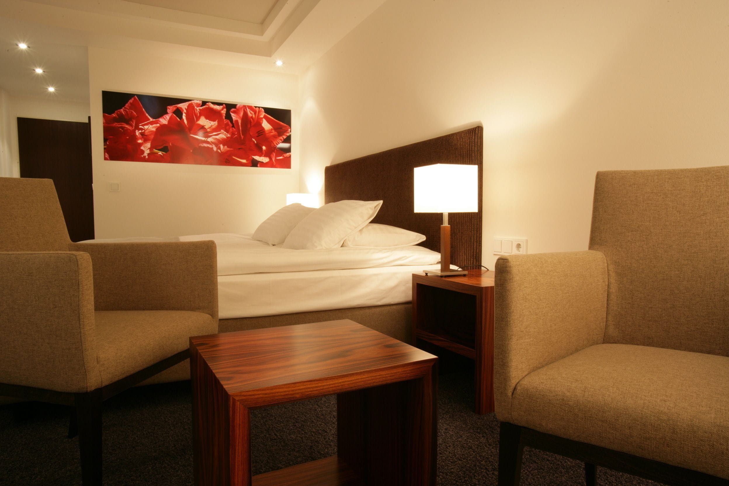 A hotel room with a bed, two armchairs, a wooden side table, and a painting on the wall.