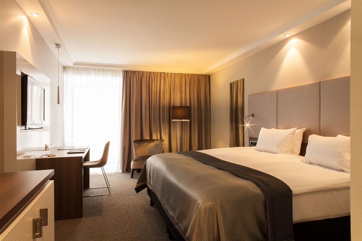 Une chambre d’hôtel avec un lit, un bureau, une chaise et une grande fenêtre avec des rideaux.