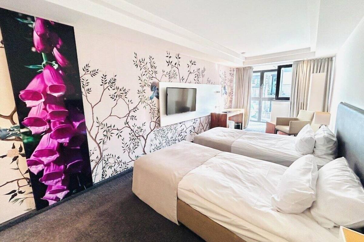 Une chambre d’hôtel avec deux lits, un téléviseur fixé au mur, un bureau avec une chaise et une grande fenêtre avec des rideaux.