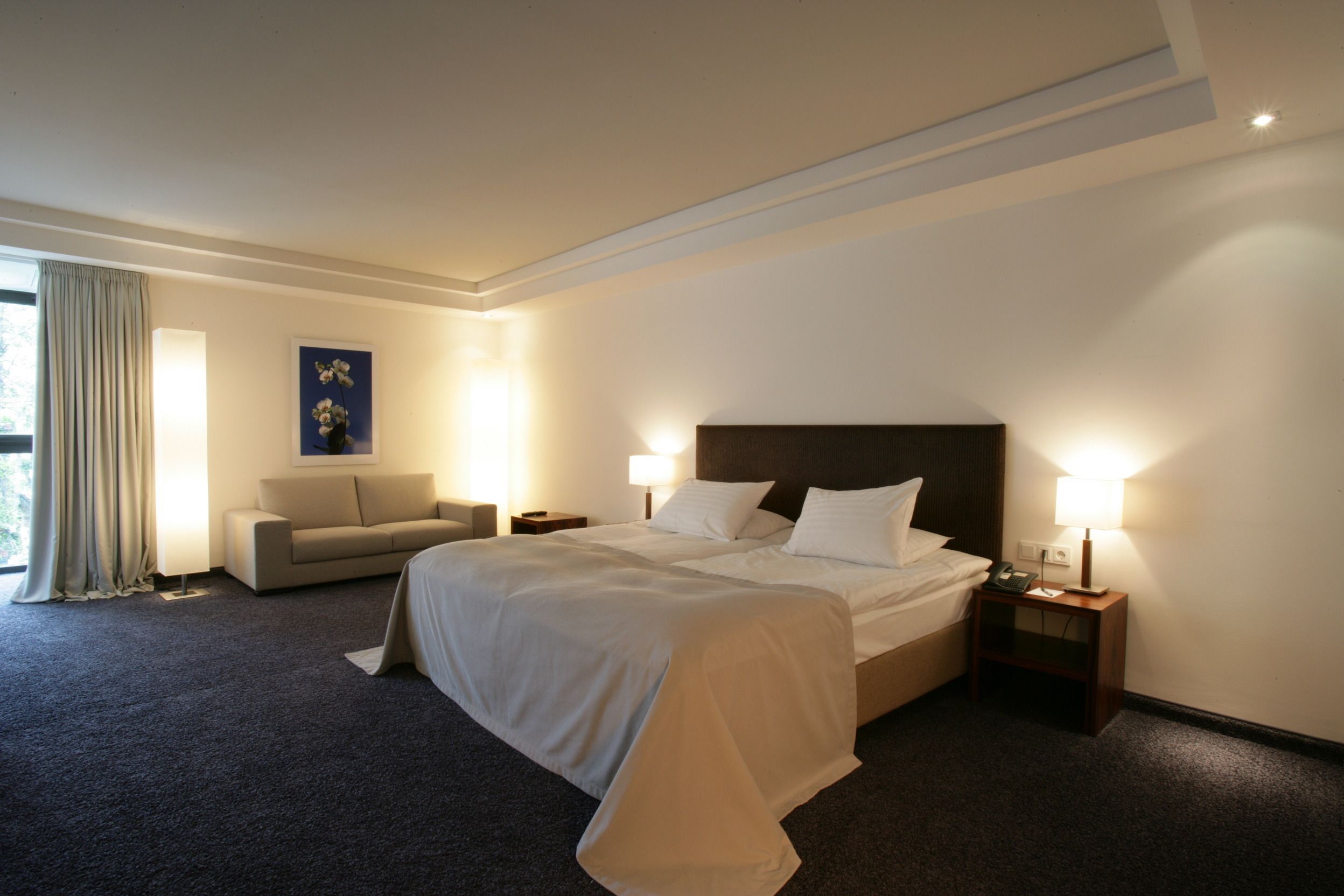 Habitación de hotel con una cama grande, un sofá, dos mesitas de noche con lámparas y un cuadro en la pared.