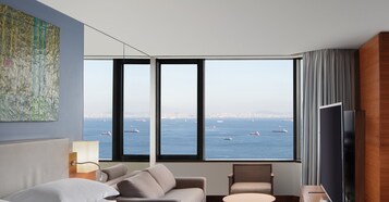Un moderno cuarto de hotel con un gran ventanal que ofrece vista a la ciudad y al océano, un sofá cómodo, un televisor de pantalla plana y un cuadro en la pared.