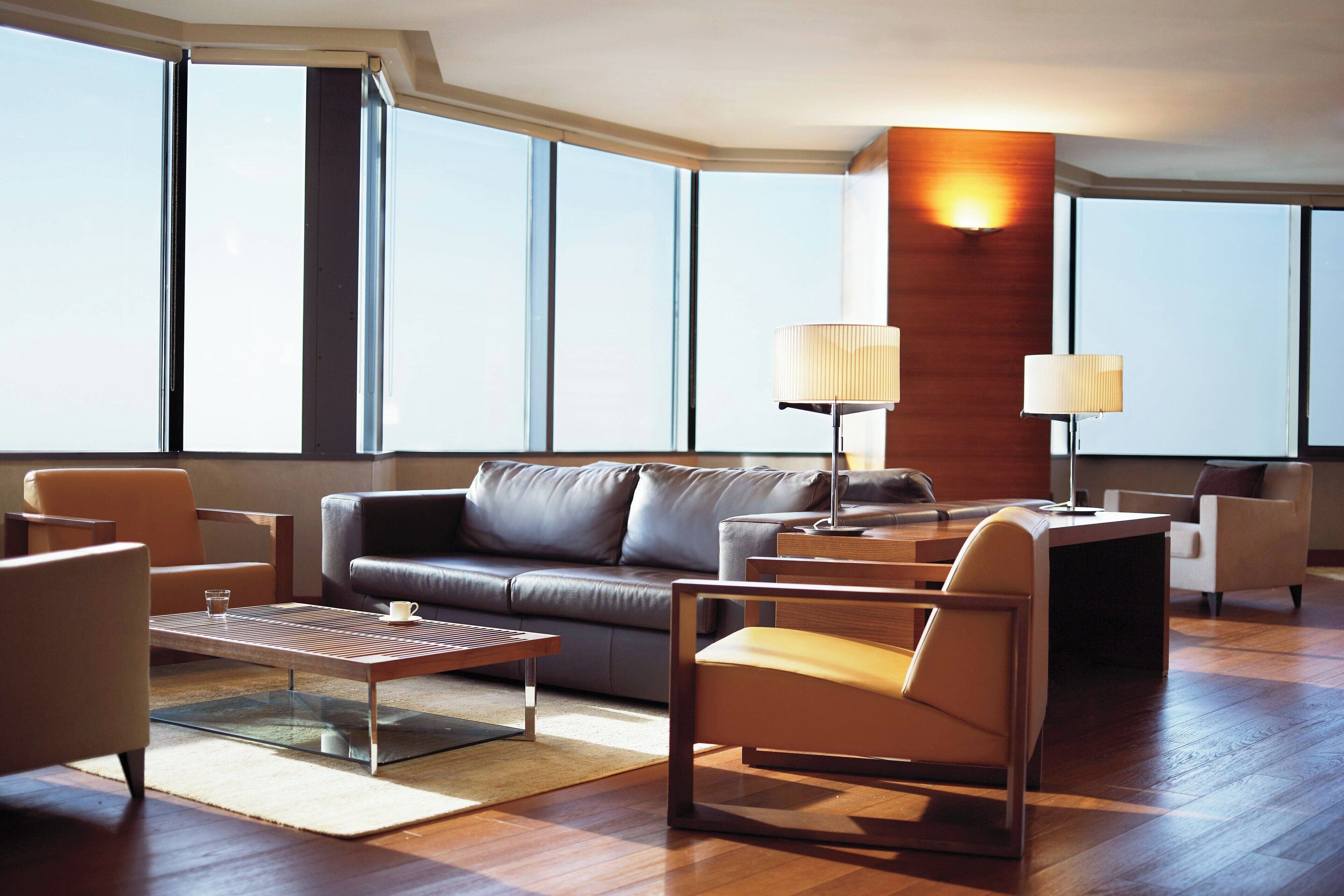 A modern living room with a leather sofa, a glass coffee table, and two lamps. Large windows provide natural light and a view of the city.