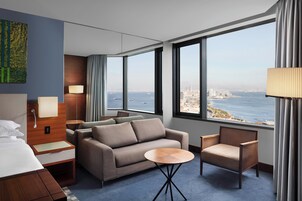 Quarto de hotel com cama, sofá, cadeira, mesinha e uma grande janela com vista para a cidade e o mar.