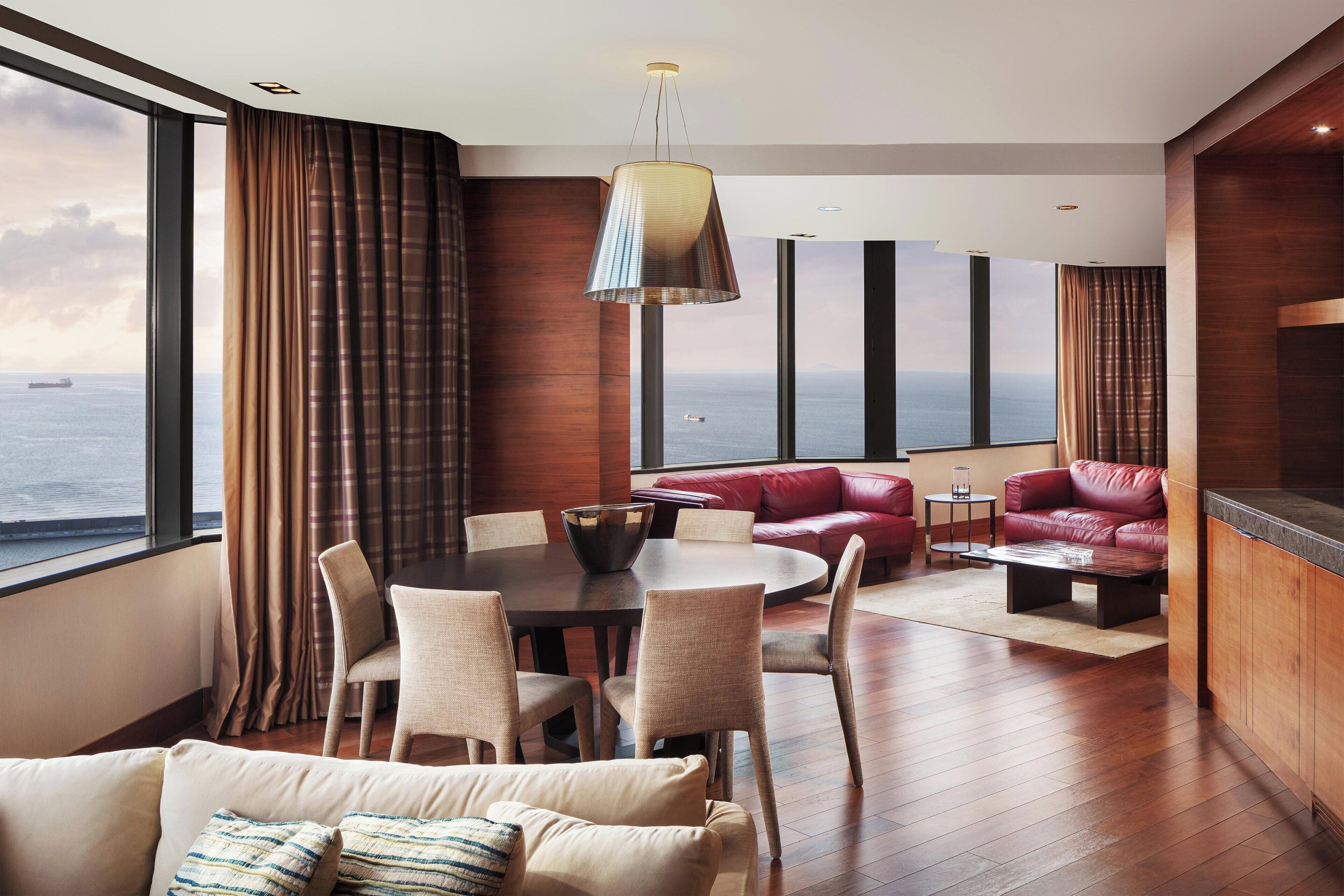 Una habitación de hotel moderna con un amplio ventanal que ofrece vistas al mar, un área de comedor con una mesa redonda y sillas, un sofá de cuero rojo y suelo de madera.