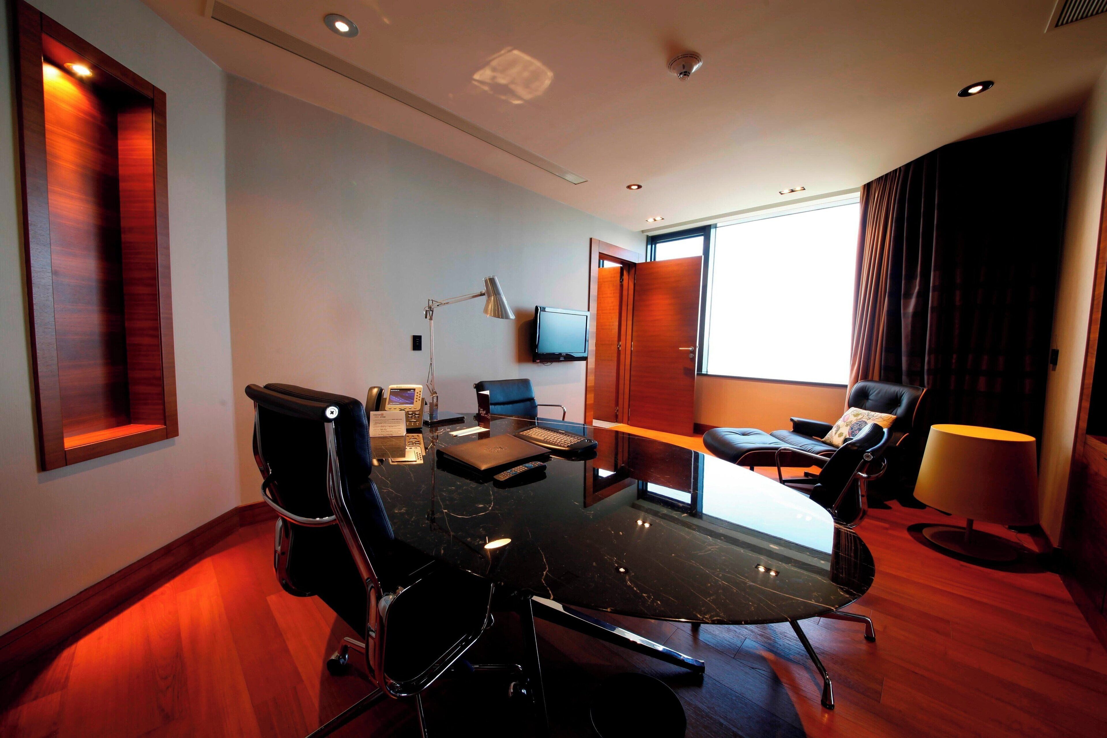 Una sala de conferencias moderna con una mesa de vidrio, sillas negras, un escritorio con computadora, una lámpara y un televisor en la pared.