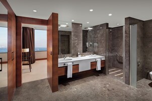 Un baño moderno con tocador de doble lavamanos, cabina de ducha con mampara de vidrio y vistas a la ciudad.
