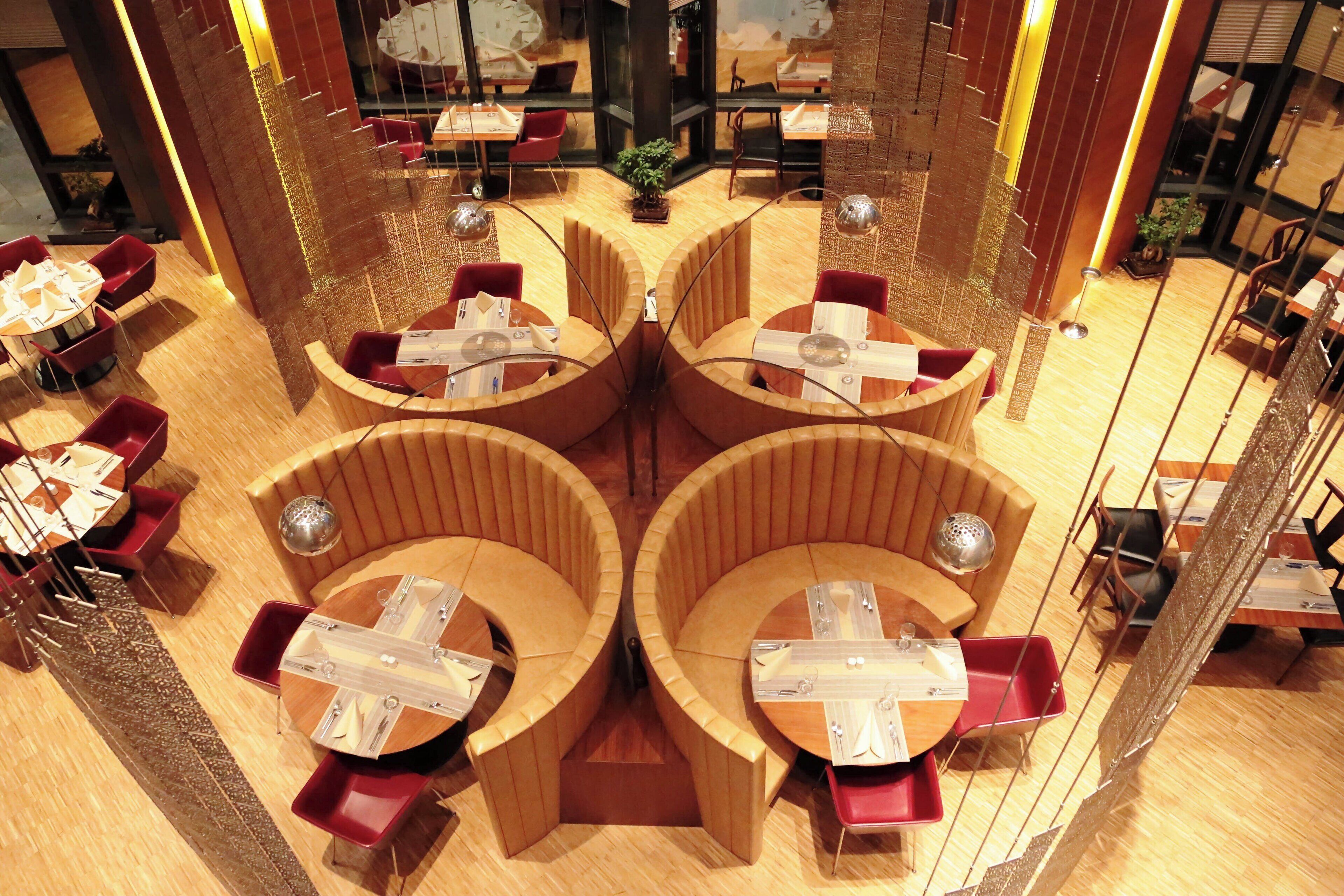 A modern restaurant with a central seating area featuring curved wooden booths and red chairs. Tables are set with white tablecloths and silverware.