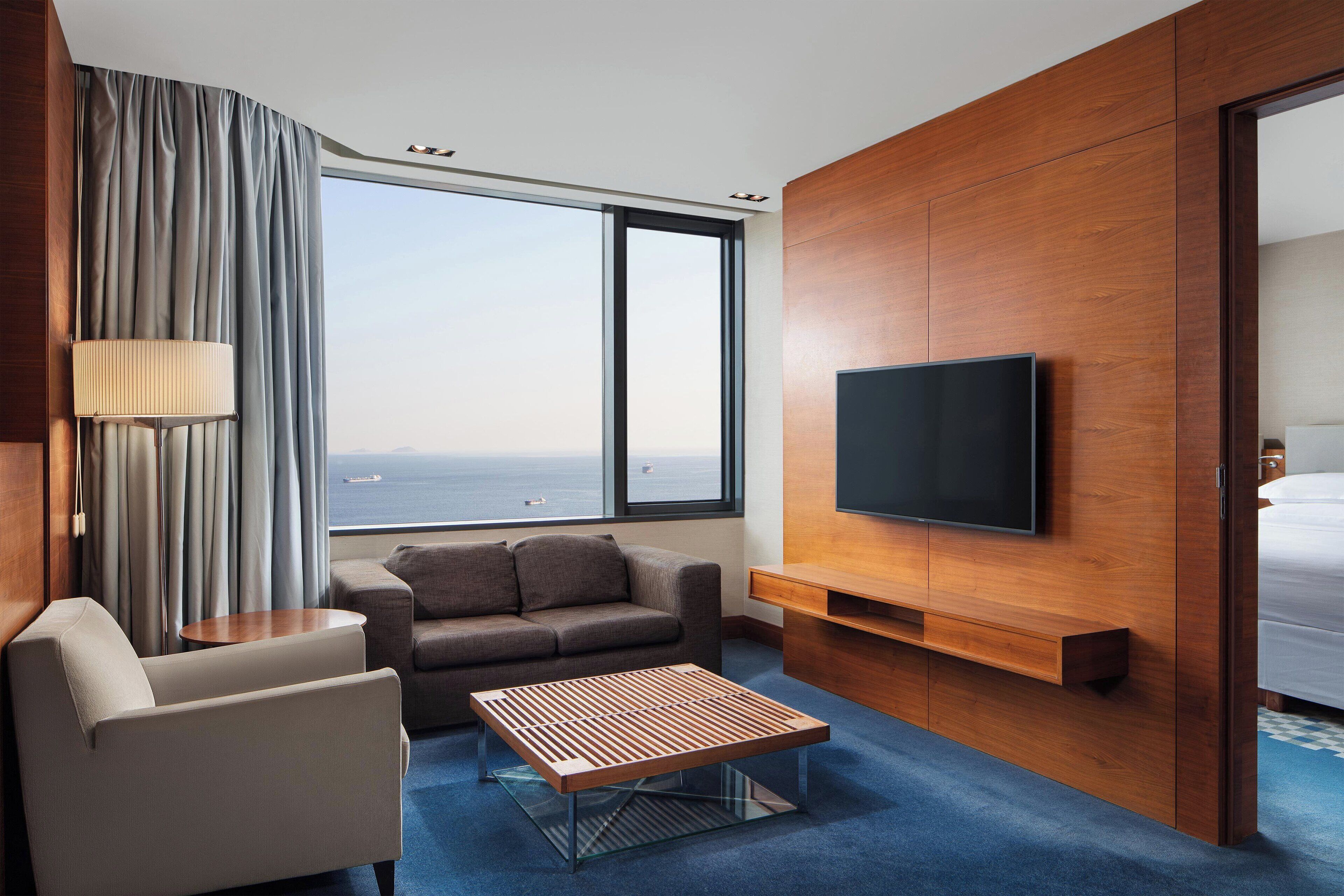 A modern hotel room with a large window, a flat-screen TV, a sofa, a coffee table, and a lamp.