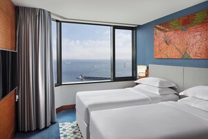 Una habitación de hotel con dos camas, un ventanal con vistas al mar y un cuadro en la pared.