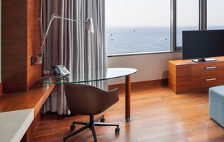 Una habitación de hotel moderna con un escritorio de madera, una mesa de vidrio, una silla, un televisor y vistas al mar a través de amplias ventanas.