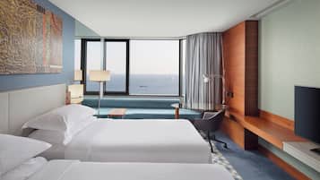 A hotel room with two beds, a desk, a chair, a TV, and a large window with a view of the sea.