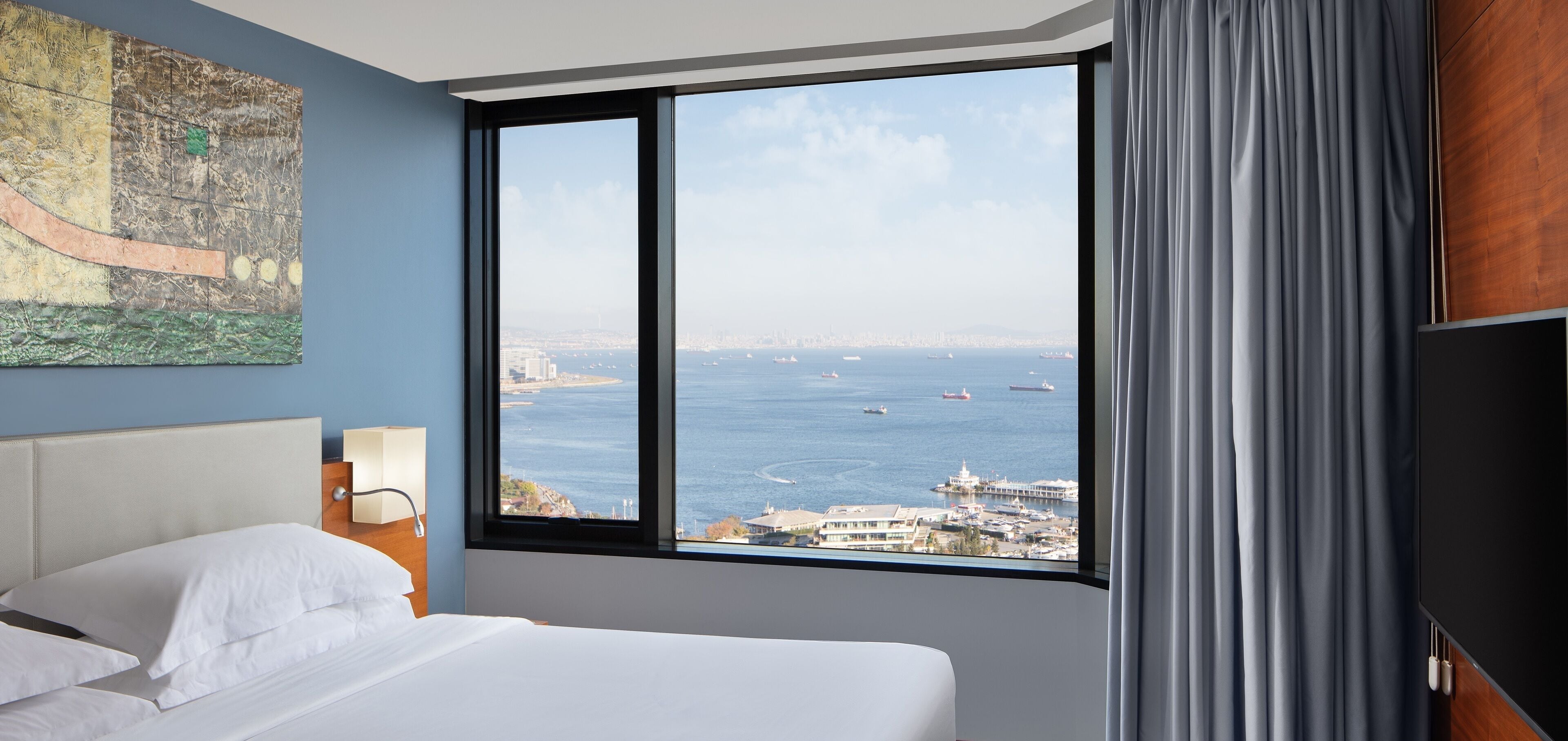 A hotel room with a large window offering a view of the sea and cityscape.