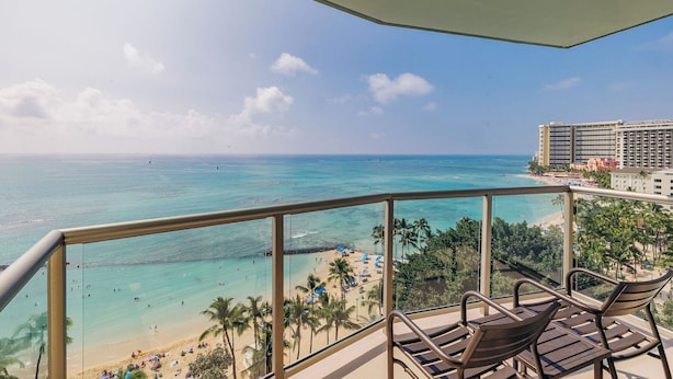 A balcony with a view of a beach and ocean, equipped with wooden chairs and a glass railing.