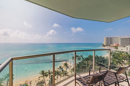 A balcony with a view of a beach and ocean, equipped with wooden chairs and a glass railing.