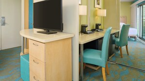 A hotel room with a desk, a flat-screen TV, a mirror, a bed, and a window.
