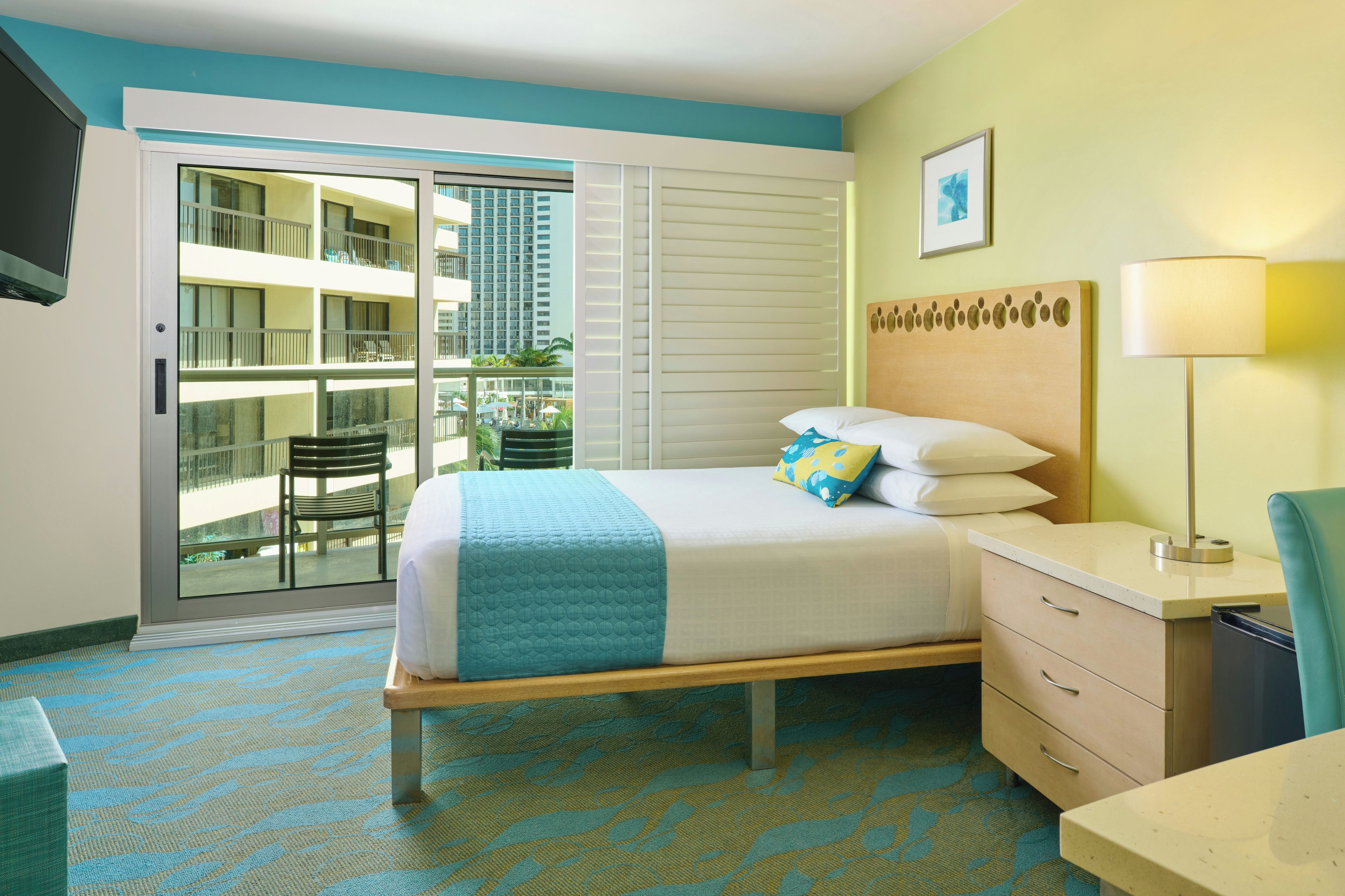 A hotel room with a bed, a desk, a chair, a window with a view of a building, and a door leading to an outdoor area.