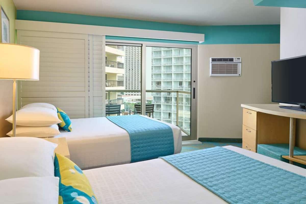 Habitación de hotel con dos camas, un escritorio, un televisor y un balcón con vista a la ciudad.