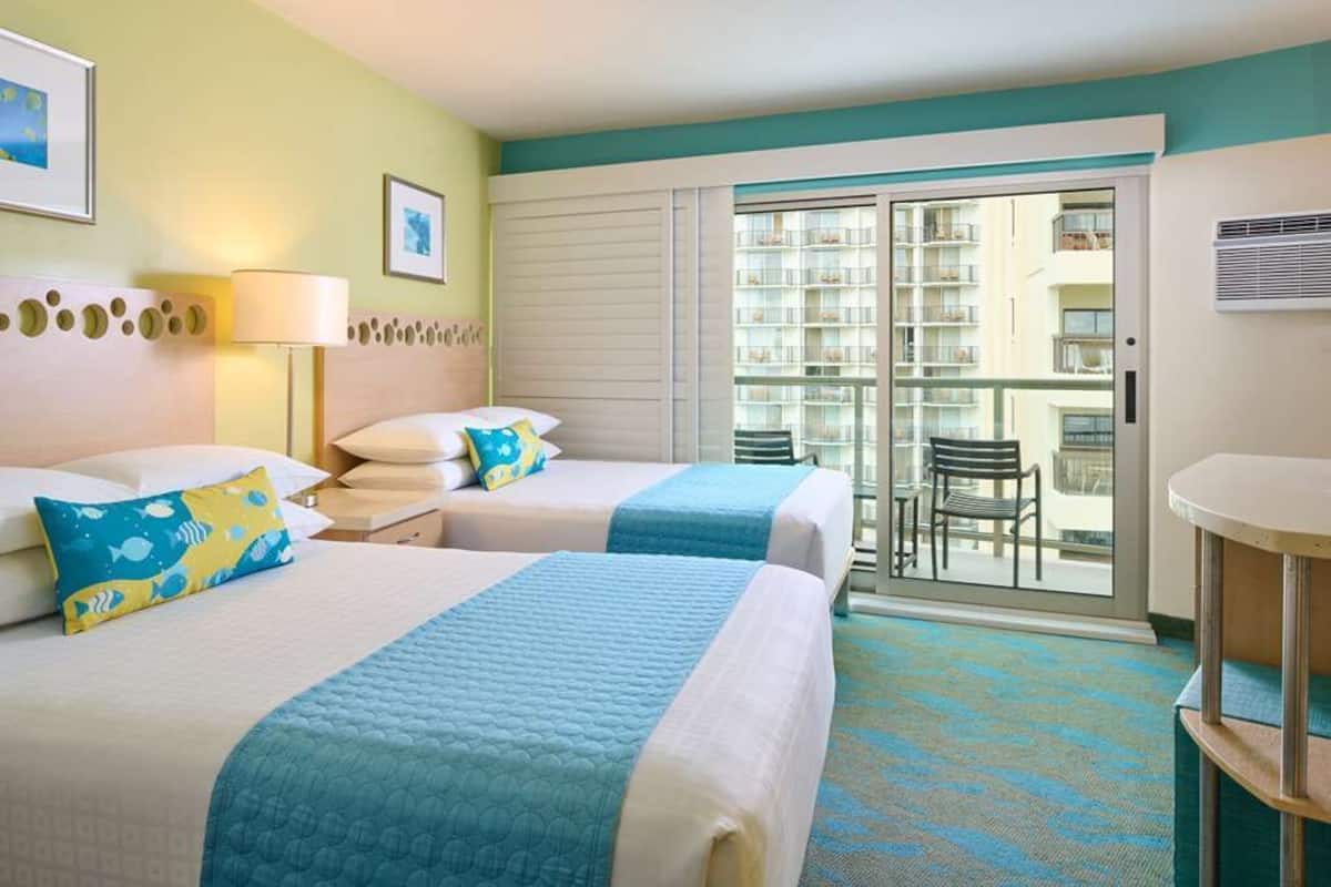 Habitación de hotel con dos camas, un balcón con vistas a otros edificios, una pequeña mesa y una silla.