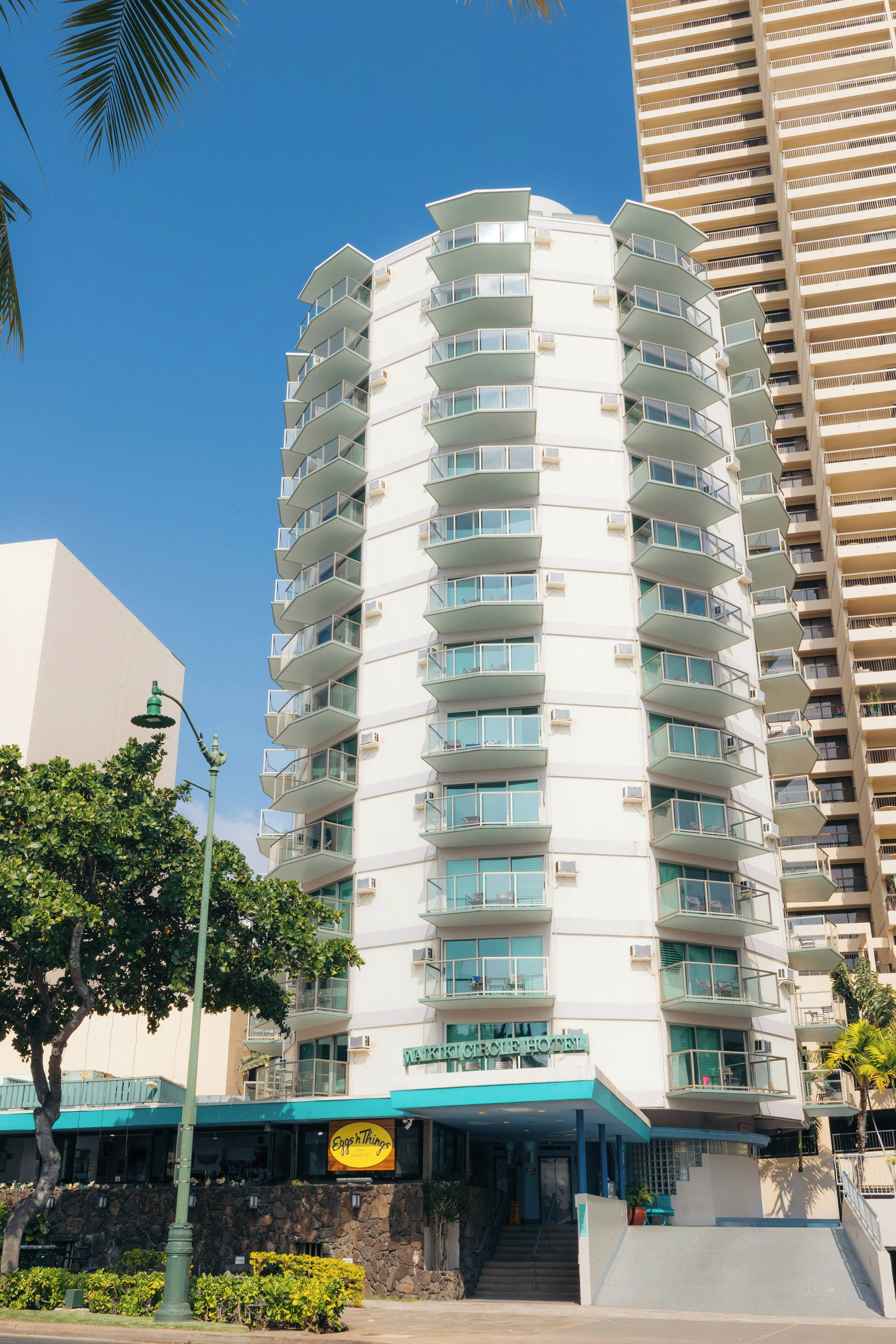 Hotel com vários andares e design moderno, com varandas em cada pavimento, fachada de pedra na base e céu azul.