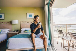 A hotel room with a bed, a bedside table with a lamp, a window with a view, and a balcony with chairs.