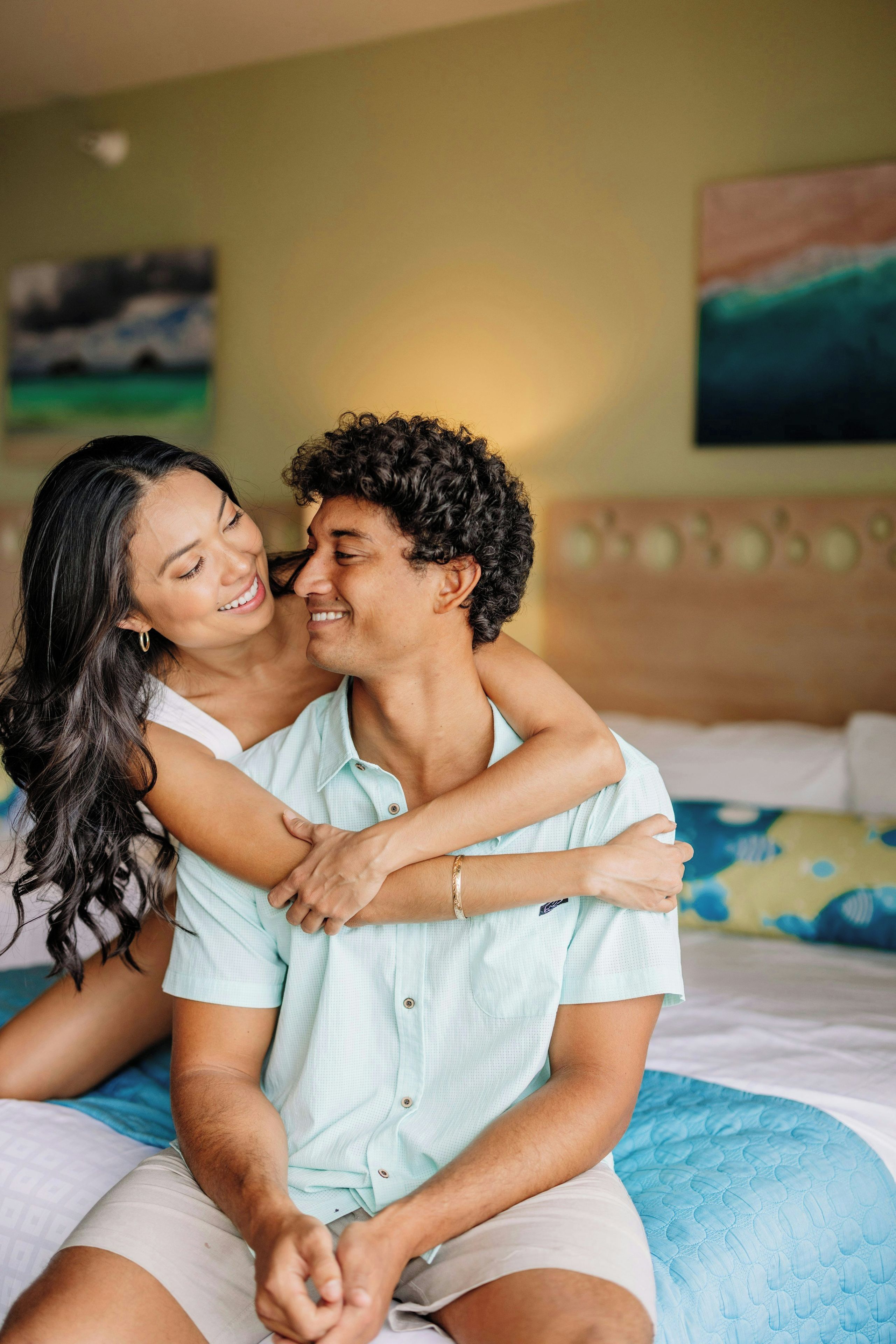 Um casal sentado na cama de um quarto de hotel, com uma parede lisa e um quadro ao fundo.