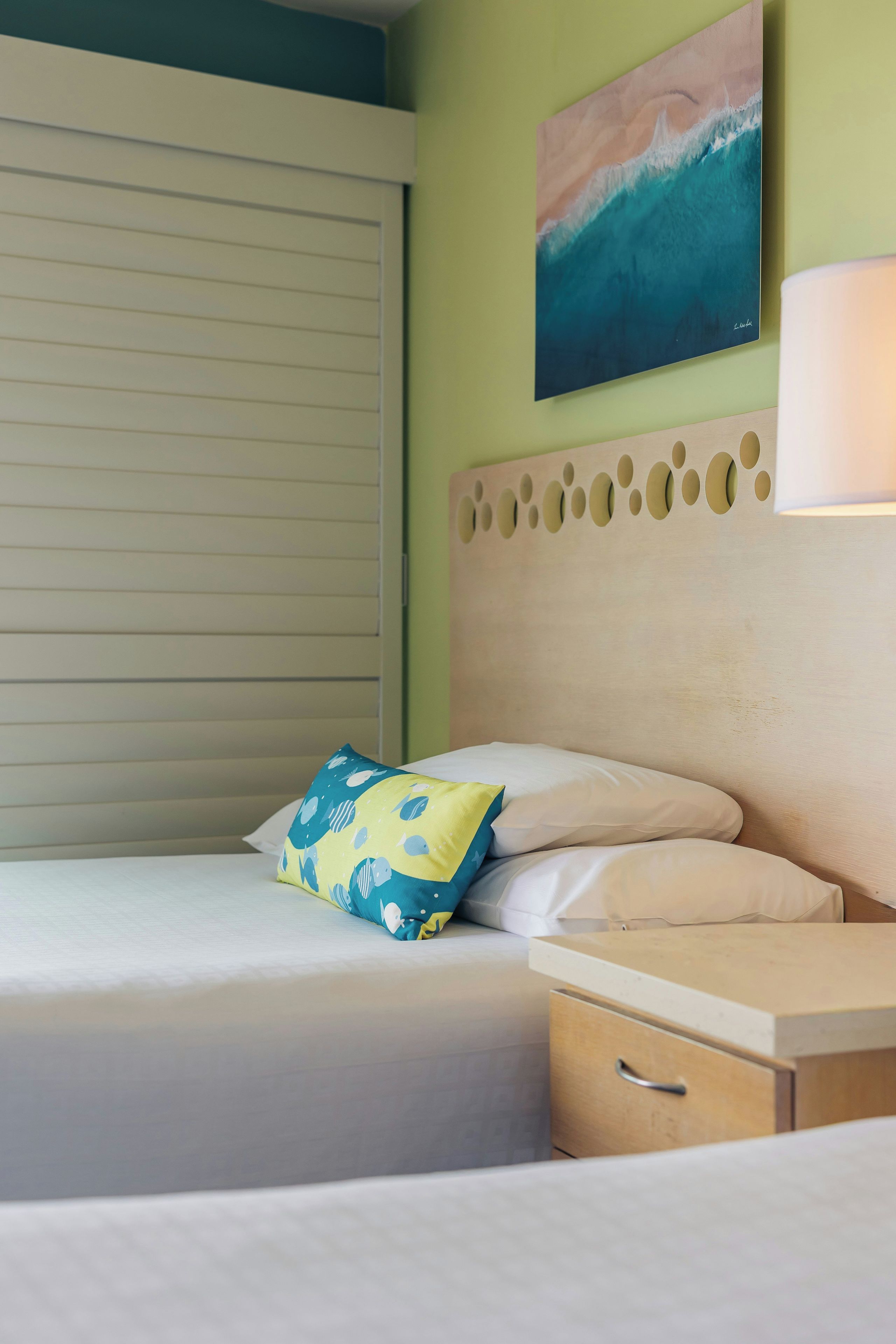 A hotel room with a bed, a nightstand, a lamp, a wall with a painting, and a window with blinds.