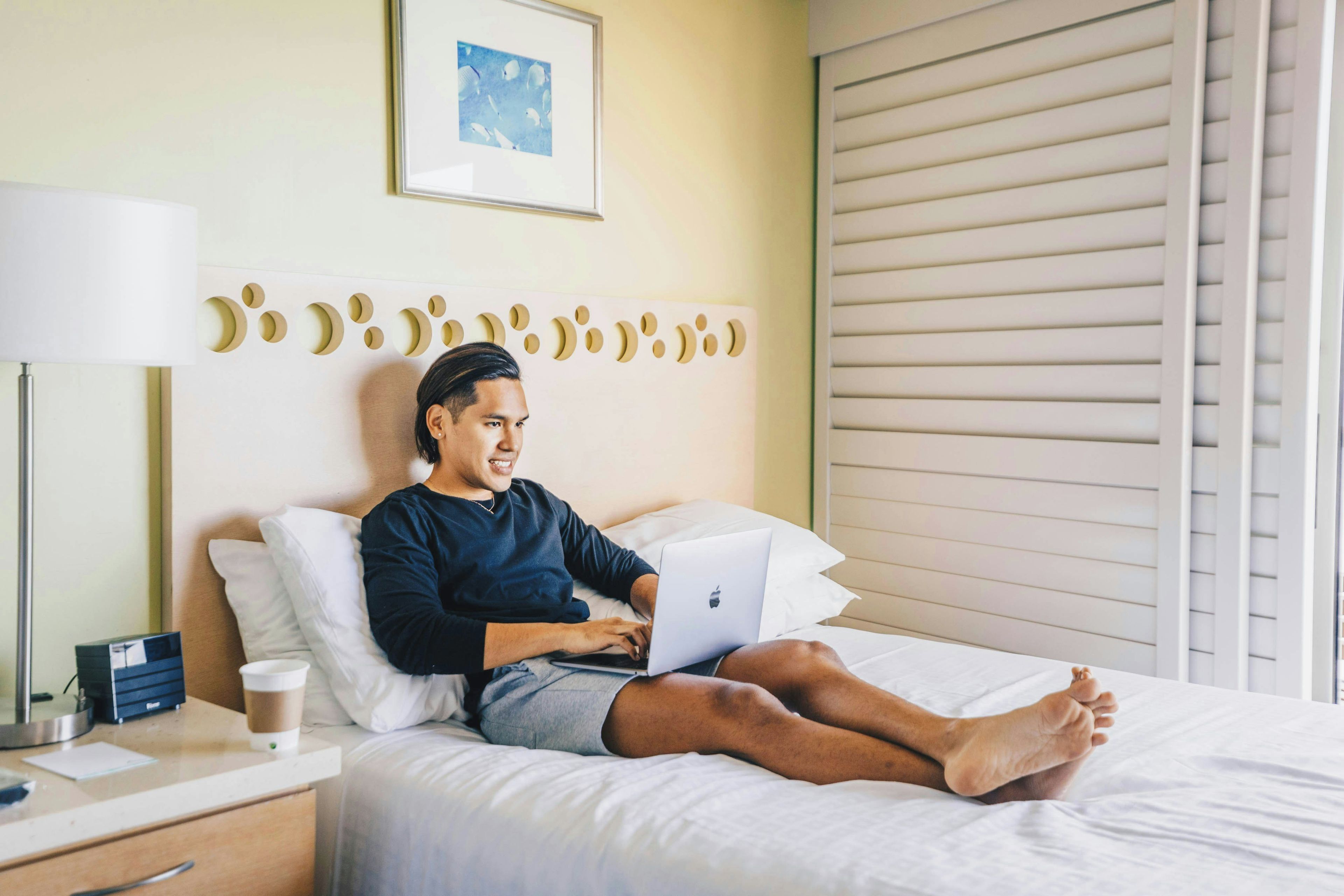 Une personne allongée sur un lit, avec un ordinateur portable, une table de chevet avec une lampe et un tableau encadré accroché au mur.