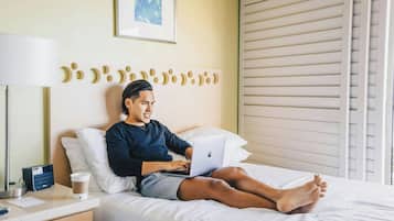 Une personne allongée sur un lit, avec un ordinateur portable, une table de chevet avec une lampe et un tableau encadré accroché au mur.