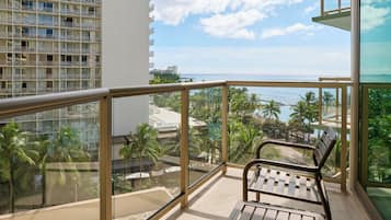 A balcony with a view of the ocean and palm trees, a chair, and a building with multiple balconies in the background.