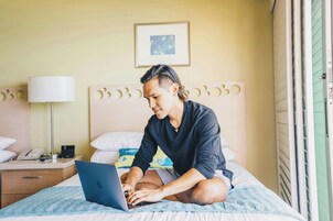 A person sitting on a bed with a laptop, a bedside table with a lamp, and a framed picture on the wall.
