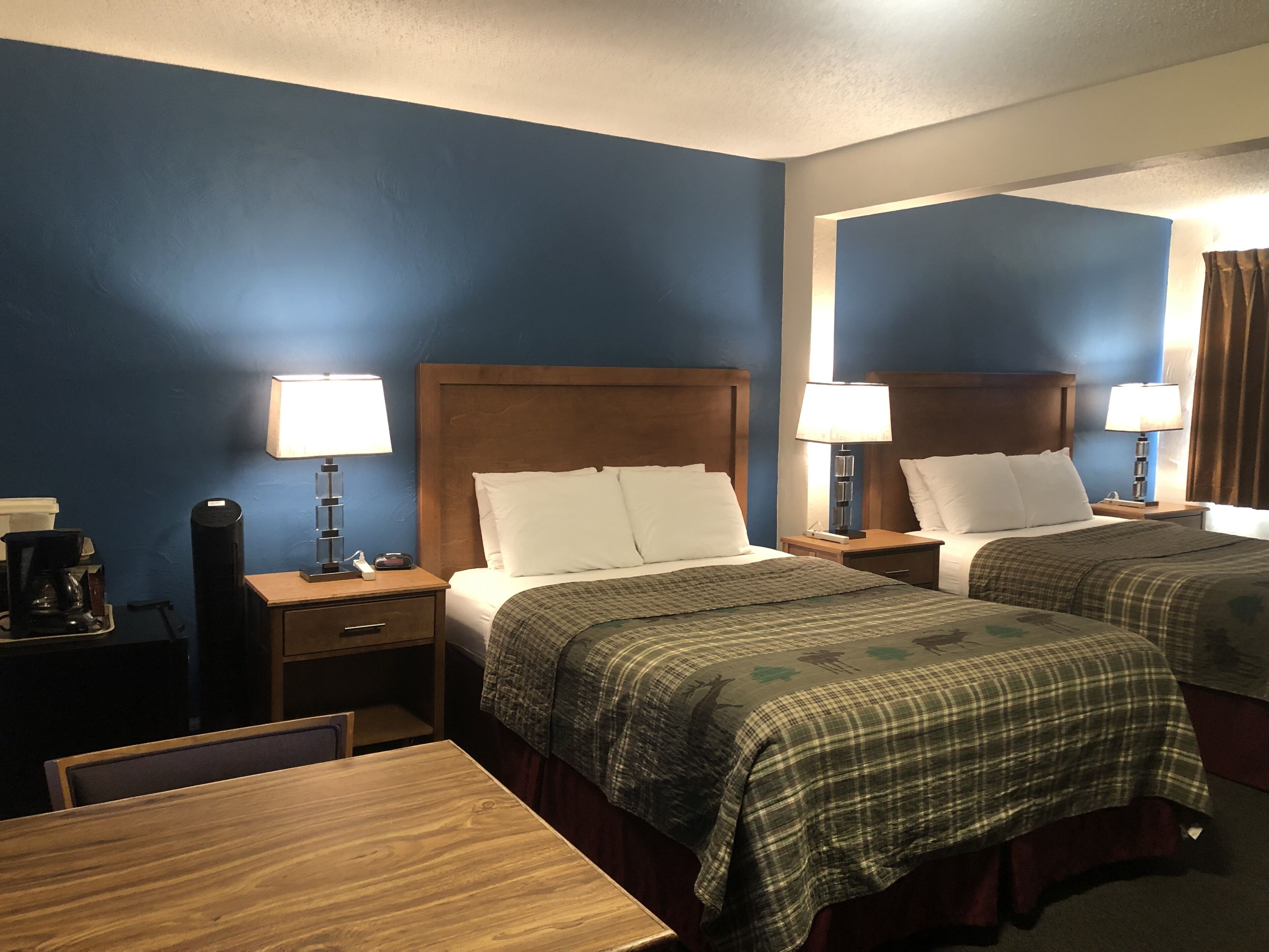 A hotel room with two beds, wooden headboards, and bedside tables with lamps. There is a wooden table and a coffee maker on the left side.