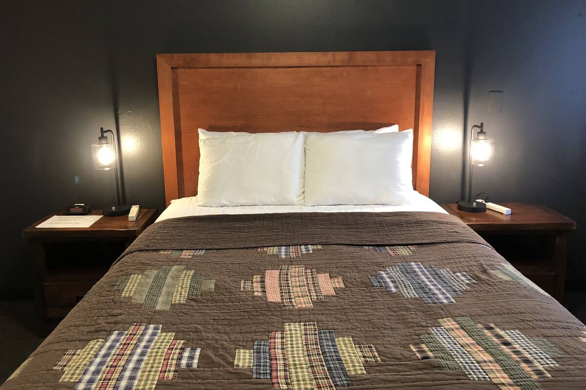 A bed with a brown quilt, white pillows, and a wooden headboard. There are two bedside tables with lamps and a small card on each.
