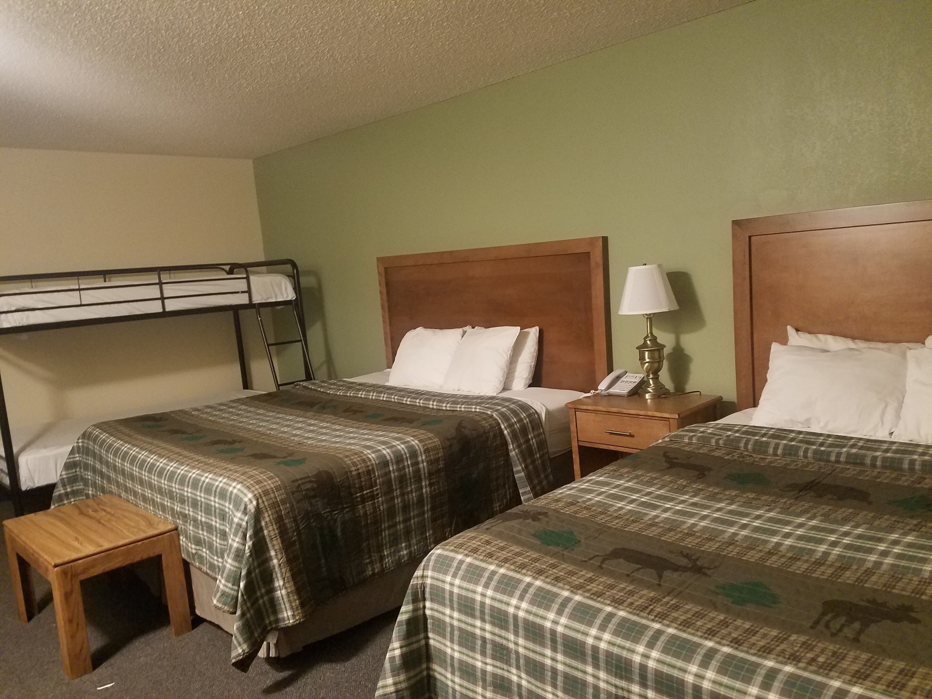 A hotel room with two beds, a bunk bed, a wooden nightstand, a lamp, and a small wooden stool.