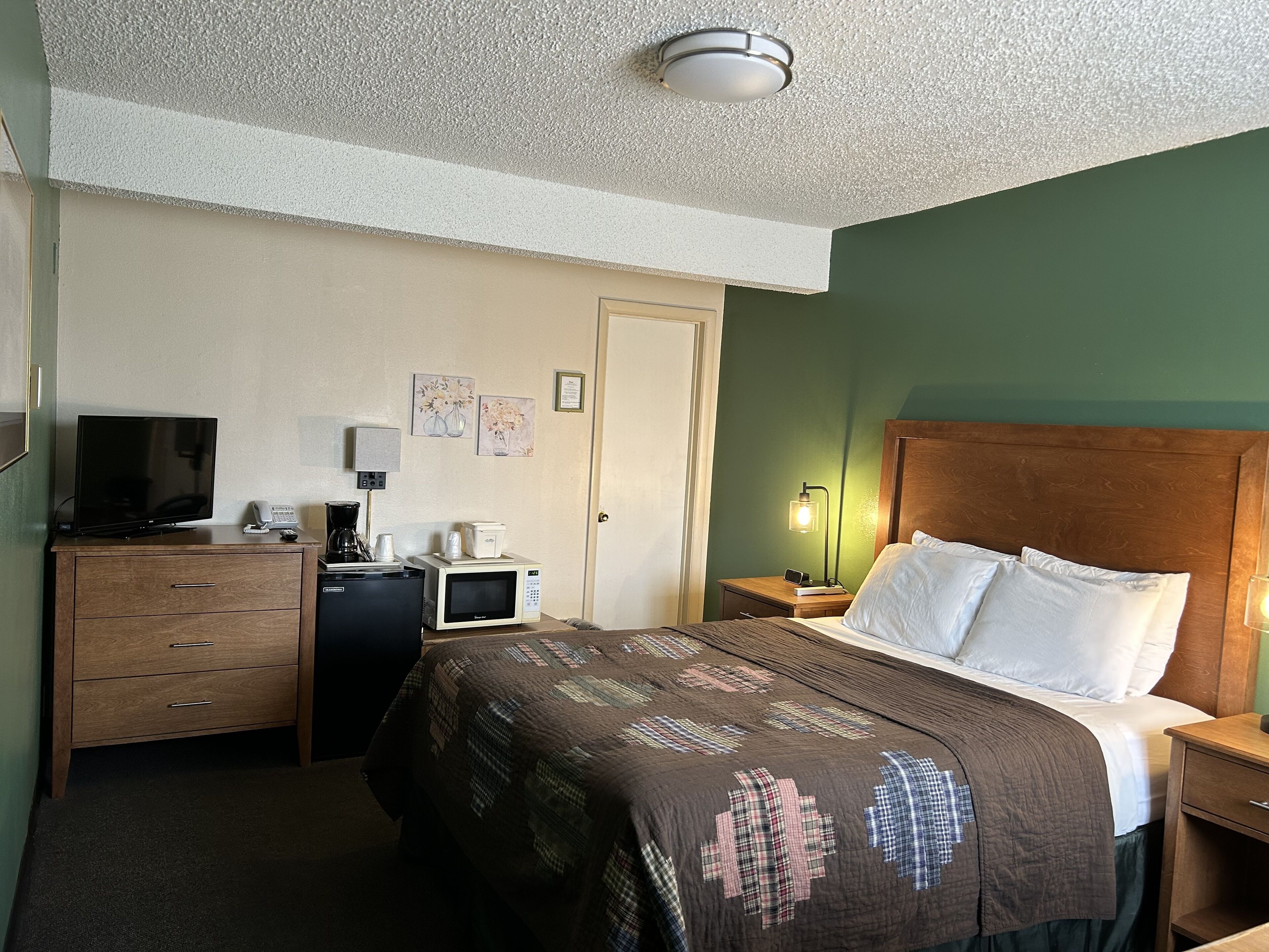 Quarto de hotel com cama, escrivaninha com televisão, micro-ondas, máquina de café e mesa de cabeceira com abajur.